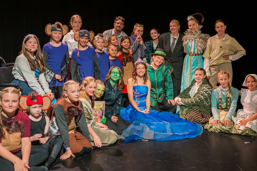 Groep kinderen en volwassenen in kleurrijke kostuums poseert op het toneel na een theatervoorstelling, met personages zoals feeën, muizen en sprookjesfiguren.