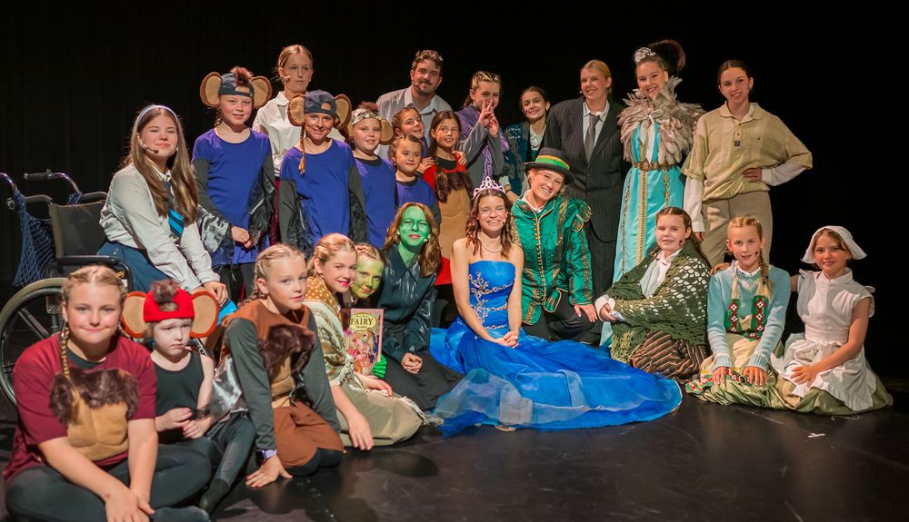 Groep kinderen en volwassenen in kleurrijke kostuums poseert op het toneel na een theatervoorstelling, met personages zoals feeën, muizen en sprookjesfiguren.