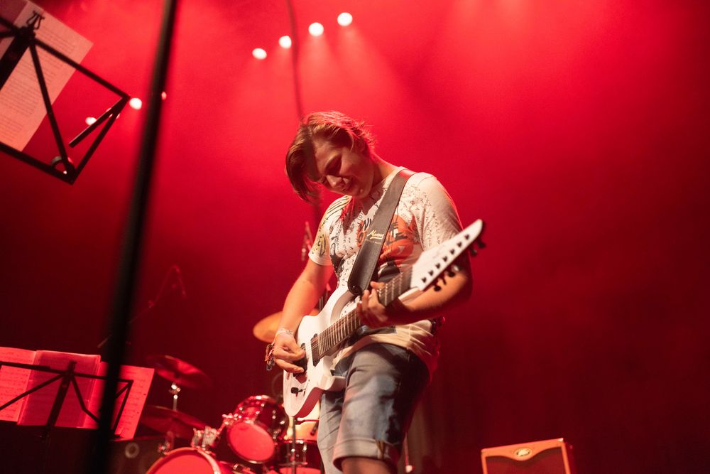 Op een podium tijdens een optreden van SCHUNCK Academy staat een muzikant onder fel rood licht en bespeelt een elektrische gitaar. Op de achtergrond staan een drumstel en versterkers, terwijl een muziekstandaard links in beeld zichtbaar is.
