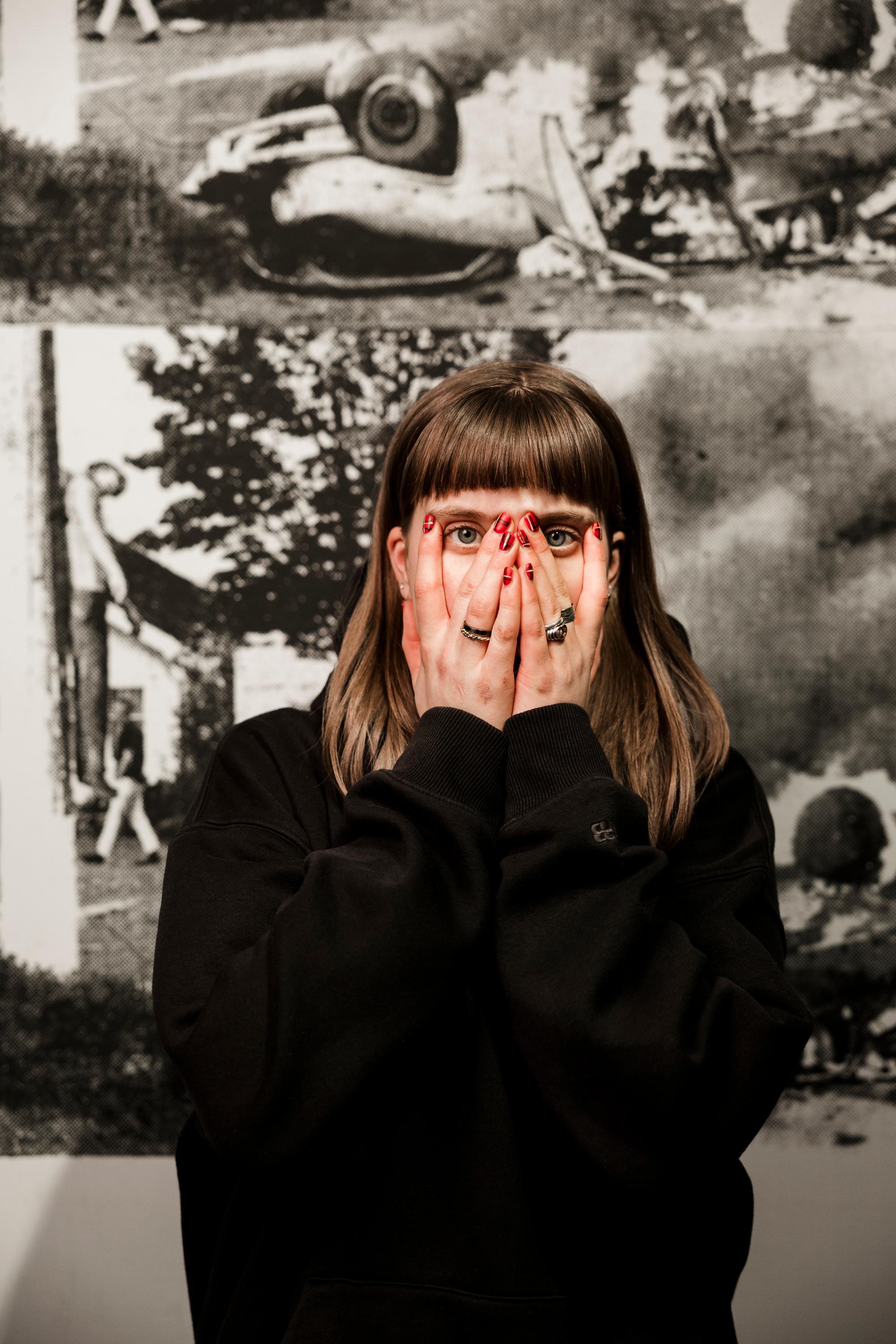 Persoon met fel rood gelakte nagels houdt handen voor mond en gezicht, terwijl ogen recht in de camera kijken; op de achtergrond een zwart-wit kunstwerk van Andy Warhol.