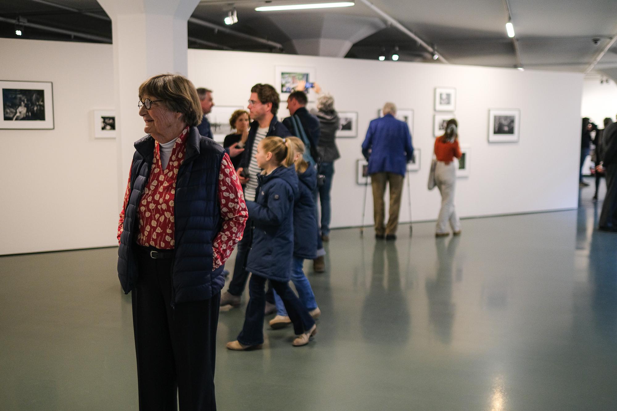 Bezoekers bekijken zwart-witfoto’s aan de wand tijdens een tentoonstelling in SCHUNCK; op de voorgrond een oudere vrouw in een rood shirt met bloemenpatroon en donker vest.