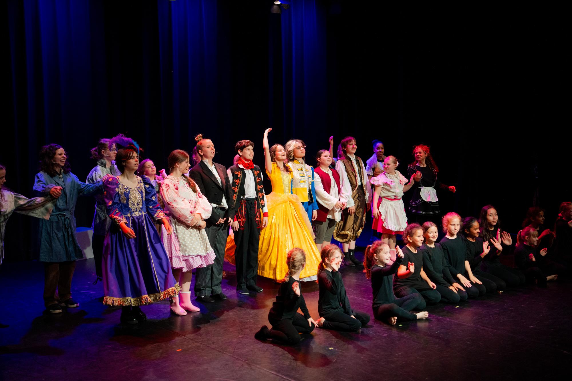Grote groep performers staat naast elkaar op een podium tijdens de musical Belle en het Beest, met in het midden een felgele jurk en daaromheen verschillende kostuums zoals jurken, pakken en themakleding. Op de voorgrond zitten enkele performers in zwarte outfits op de grond terwijl blauwe podiumverlichting en een donker gordijn de achtergrond vormen.
