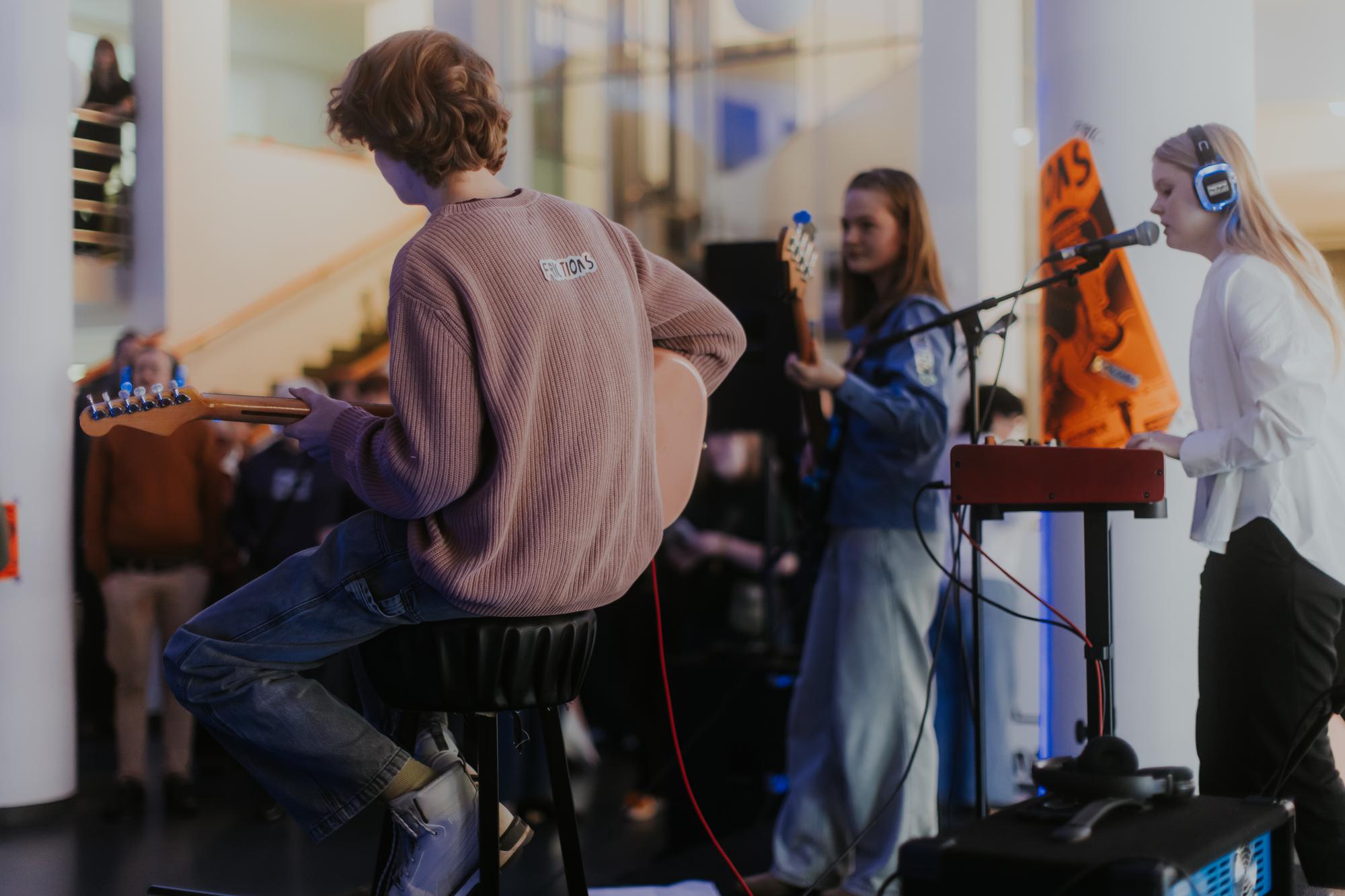 Jongerenband treedt op binnen een moderne ruimte; een jongen speelt gitaar, terwijl twee meiden basgitaar en keyboard bespelen en zingen in microfoons, met publiek op de achtergrond.
