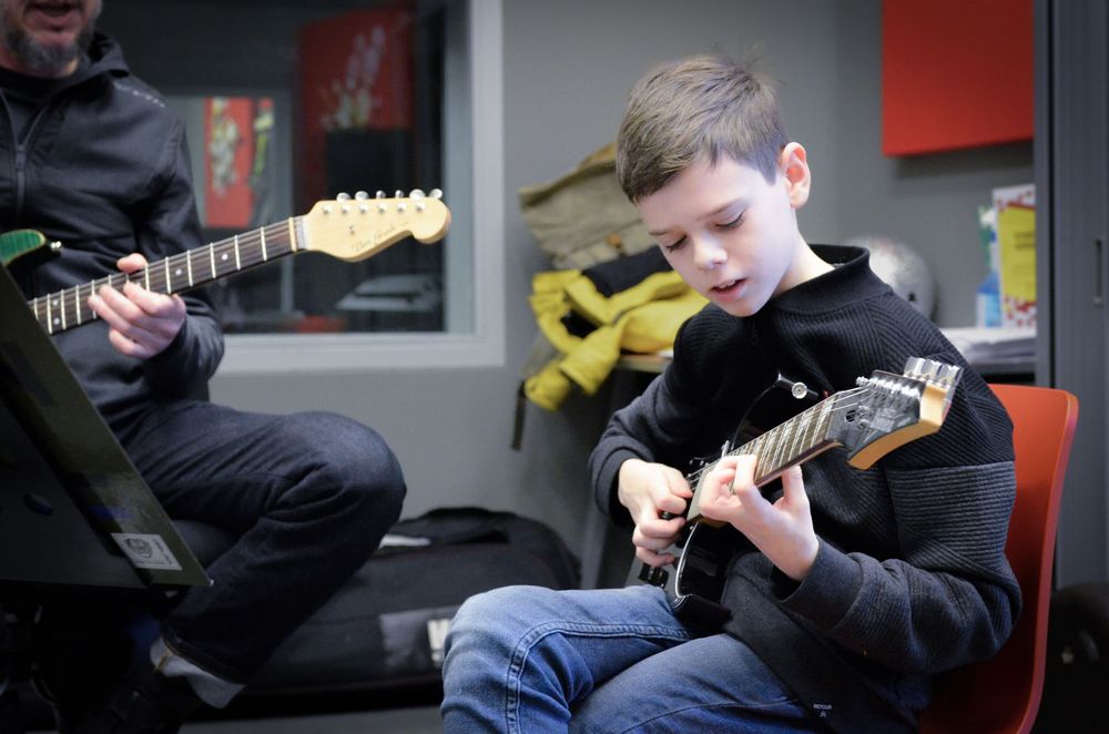 Kind zit op een stoel en speelt een elektrische gitaar tijdens een proeverij pop en rock instrumenten terwijl een volwassene naast hem ook gitaar speelt. Op de achtergrond zijn een raam, tassen en een muzieklokaal met apparatuur zichtbaar.
