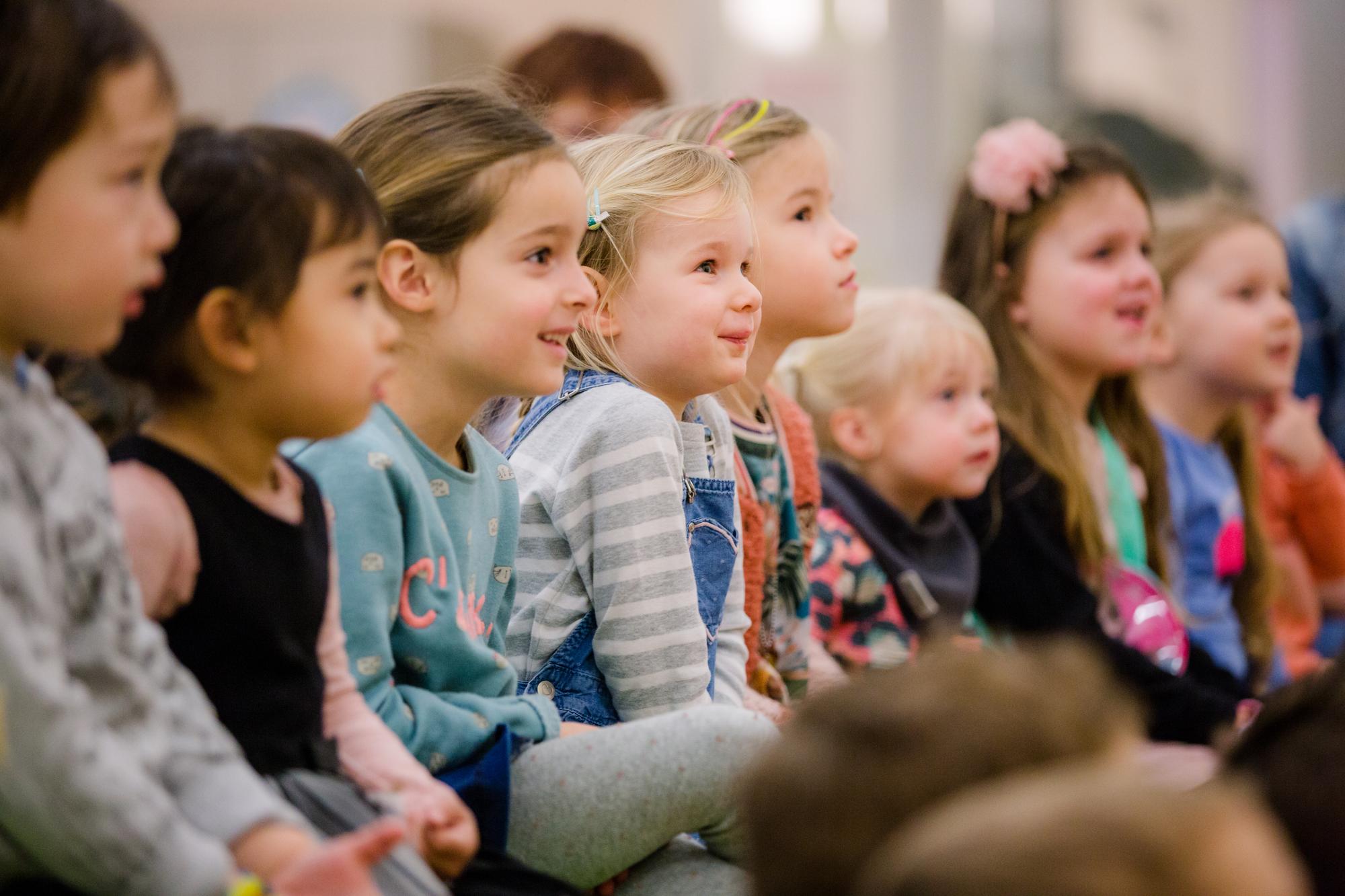 Een aantal kinderen die in een rijtje naast elkaar zitten in het publiek en hun lachende gezichtsuitdrukkingen. 