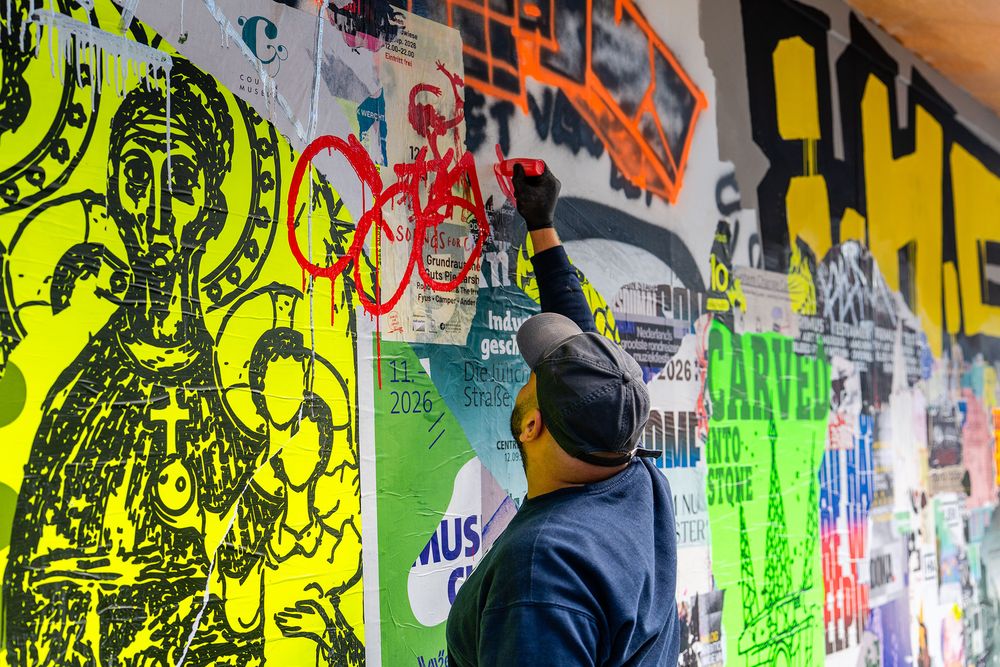 Kunstenaar Massoud Sakhi brengt met rode spuitverf graffiti aan op MODULE #6 bij het Glaspaleis in Heerlen. De muur is bedekt met kleurrijke posters, illustraties en grote gele letters.
