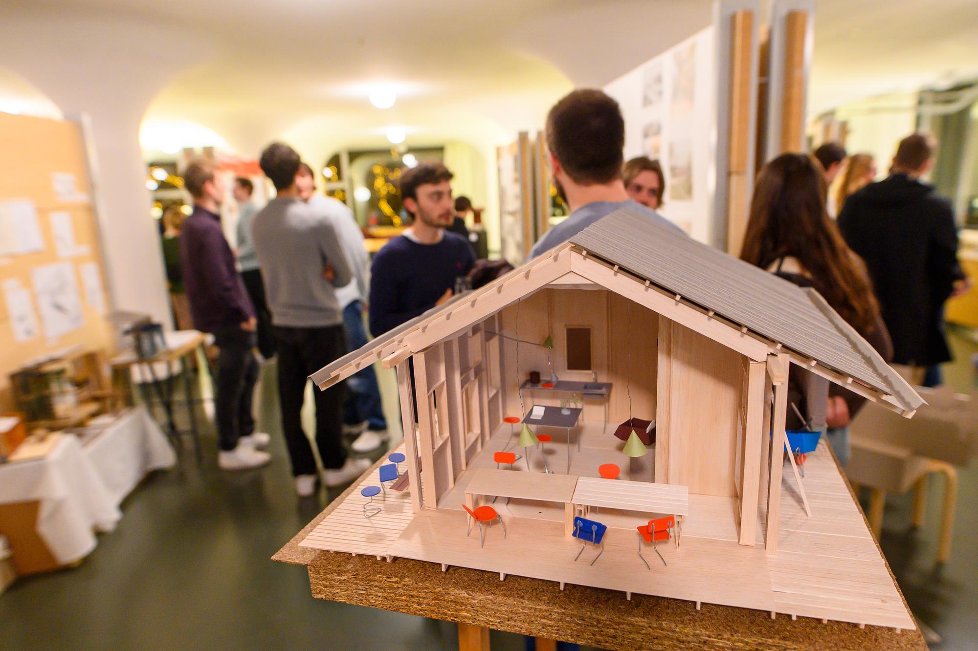 Maquette van een houten paviljoen op de voorgrond, met bezoekers die de afstudeerprojecten bekijken tijdens de tentoonstelling van de Euregional Architecture Prize in SCHUNCK.