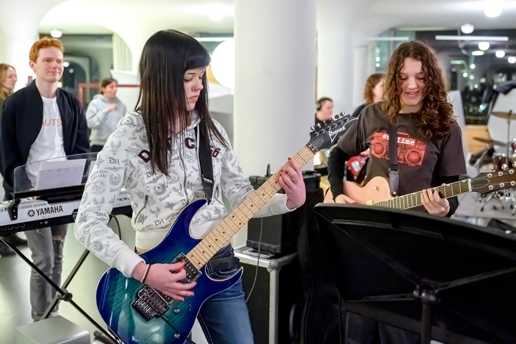 Twee meiden treden op en spelen gitaar. Achter hen speelt een jongen op een keyboard.