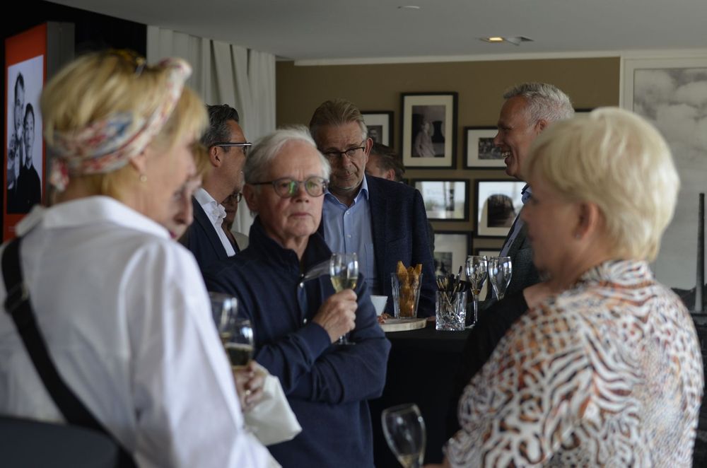 Groep volwassenen in gesprek tijdens een sponsoravond, met drankjes in de hand, in een sfeervolle ruimte met zwart-witte kunstfoto’s aan de muur.