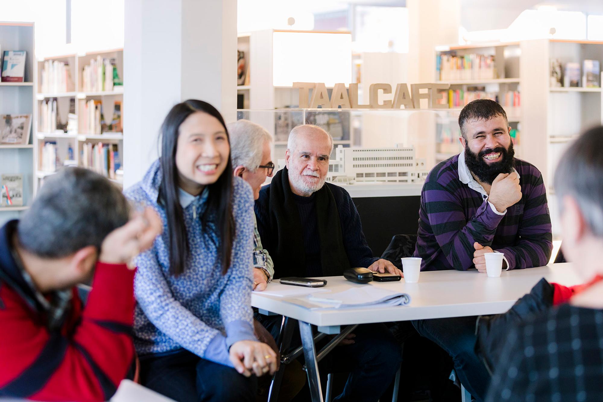 Taalcafé met zes personen die aan tafel met elkaar praten en lachen in de bibliotheek van SCHUNCK. 