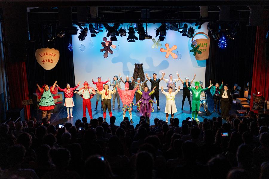 Grote groep performers in kleurrijke kostuums staat op een podium en buigt voor het publiek na een voorstelling, met decorstukken en theaterverlichting op de achtergrond.