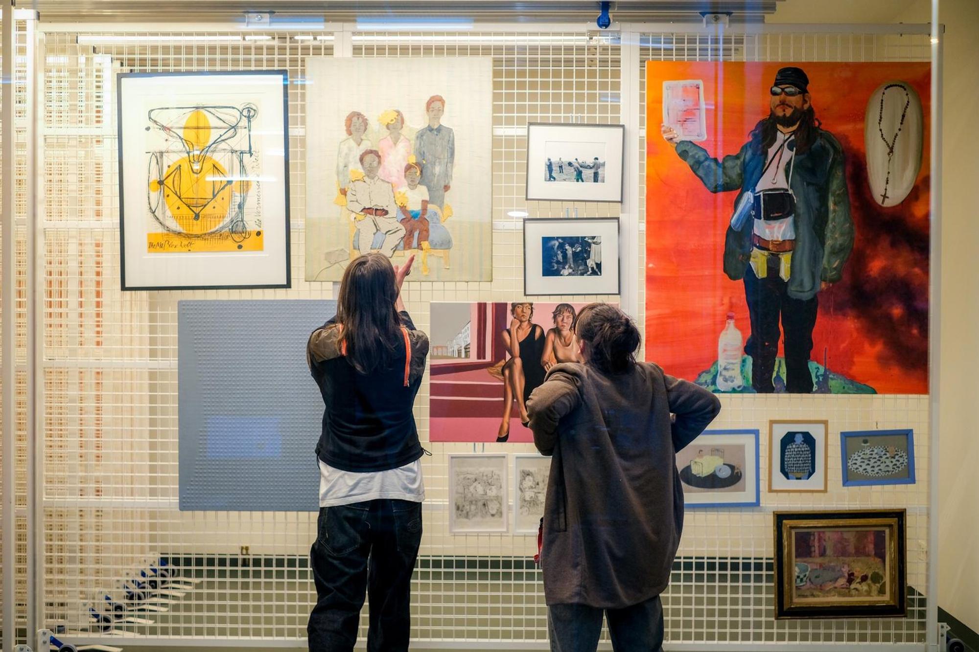 Twee personen staan in een museumruimte voor een wand met een diverse selectie kunstwerken. Ze bekijken de werken aandachtig en lijken in gesprek over de getoonde kunst. De wand bestaat uit schilderijen, tekeningen en foto’s in verschillende stijlen en formaten