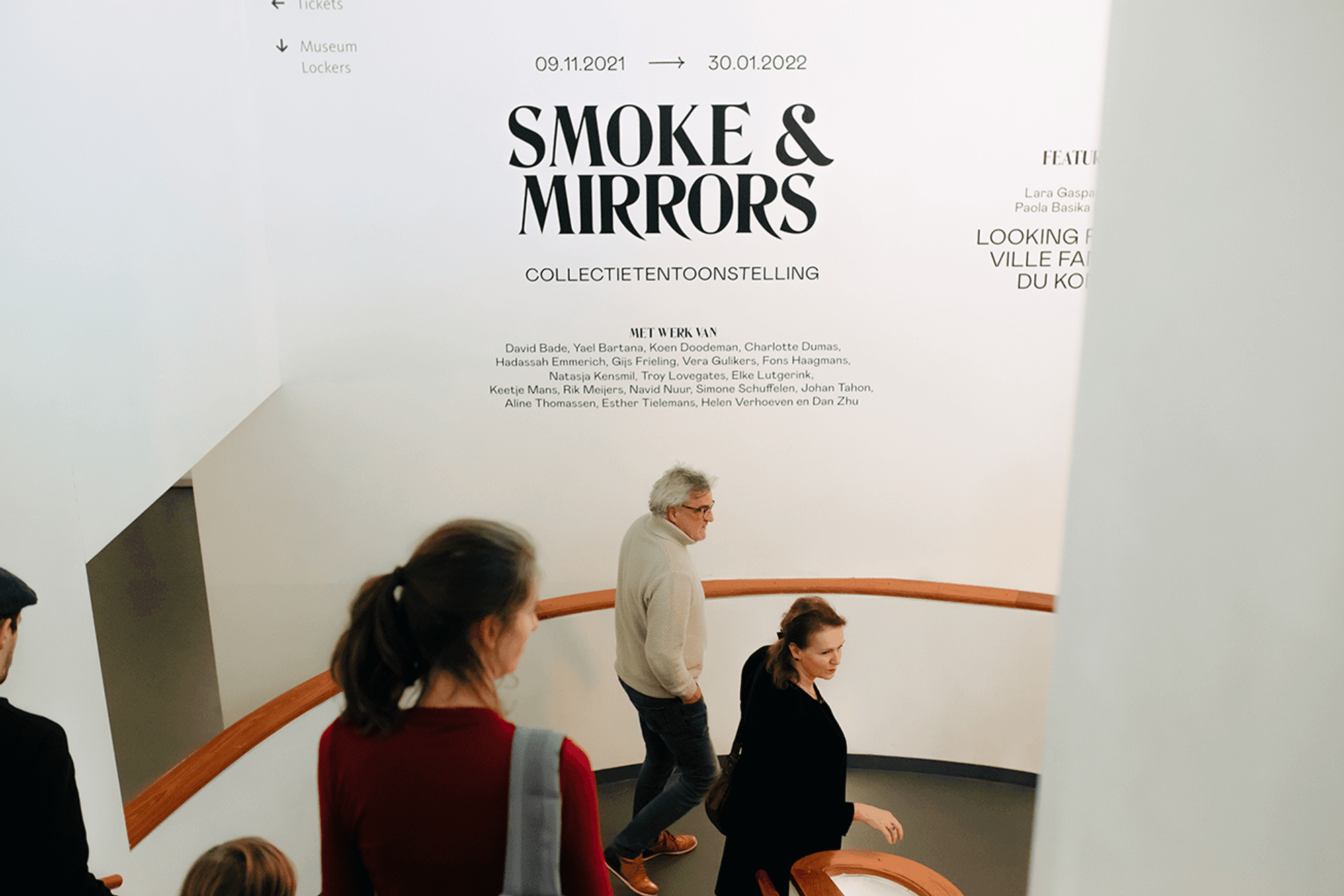 Bezoekers lopen de trap af richting het museum. Op de wand staat de titel van de tentoonstelling: 'Smoke & Mirrors'. 