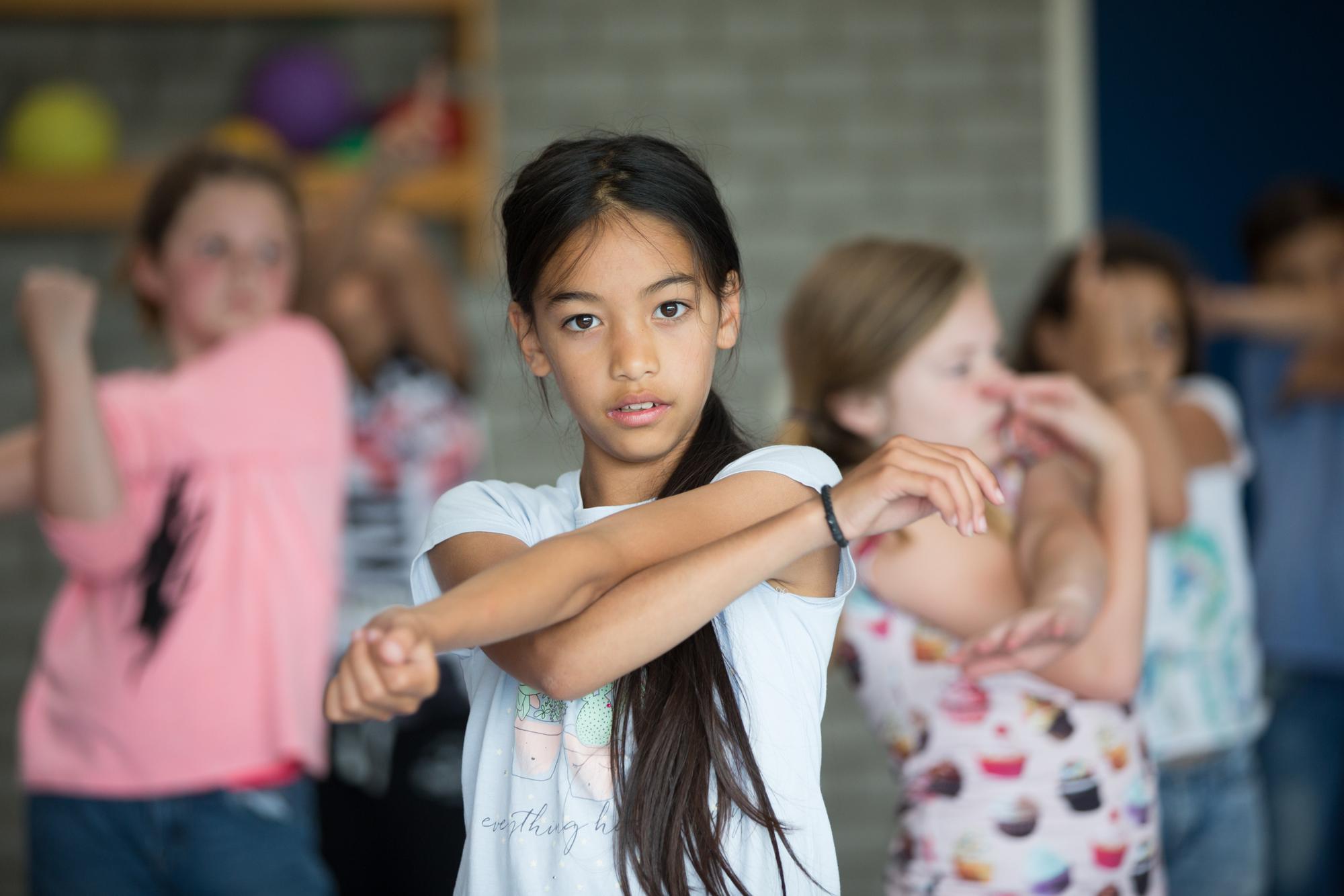 Jong meisje voert een stretchoefening uit met haar armen over elkaar, terwijl op de achtergrond meerdere andere meiden dezelfde oefening doen, onscherp in beeld.
