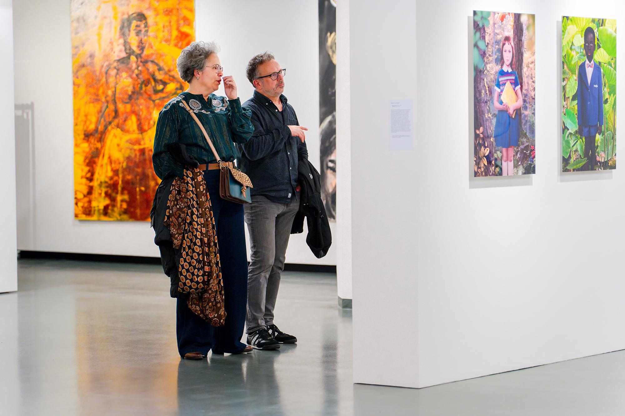 Twee personen staan in een museumruimte en bekijken ingelijste kunstwerken aan een witte wand; één persoon wijst naar een werk.