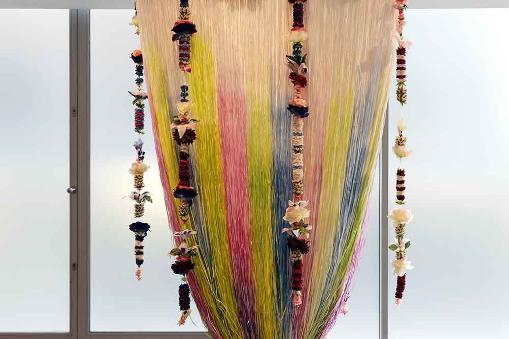 Gekleurde draden in pastel- en regenboogtinten hangen in een sierlijke boog naar beneden. Ertussen bungelen bloemrijke ornamenten, wat het geheel een feestelijk en speels karakter geeft.