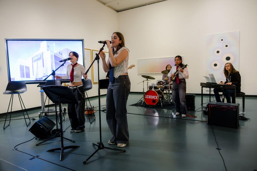 SCHUNCK Academy, jonge band treedt op in een museumruimte met zang, gitaar, drums en keyboard, tussen kunstwerken en een projectiescherm.