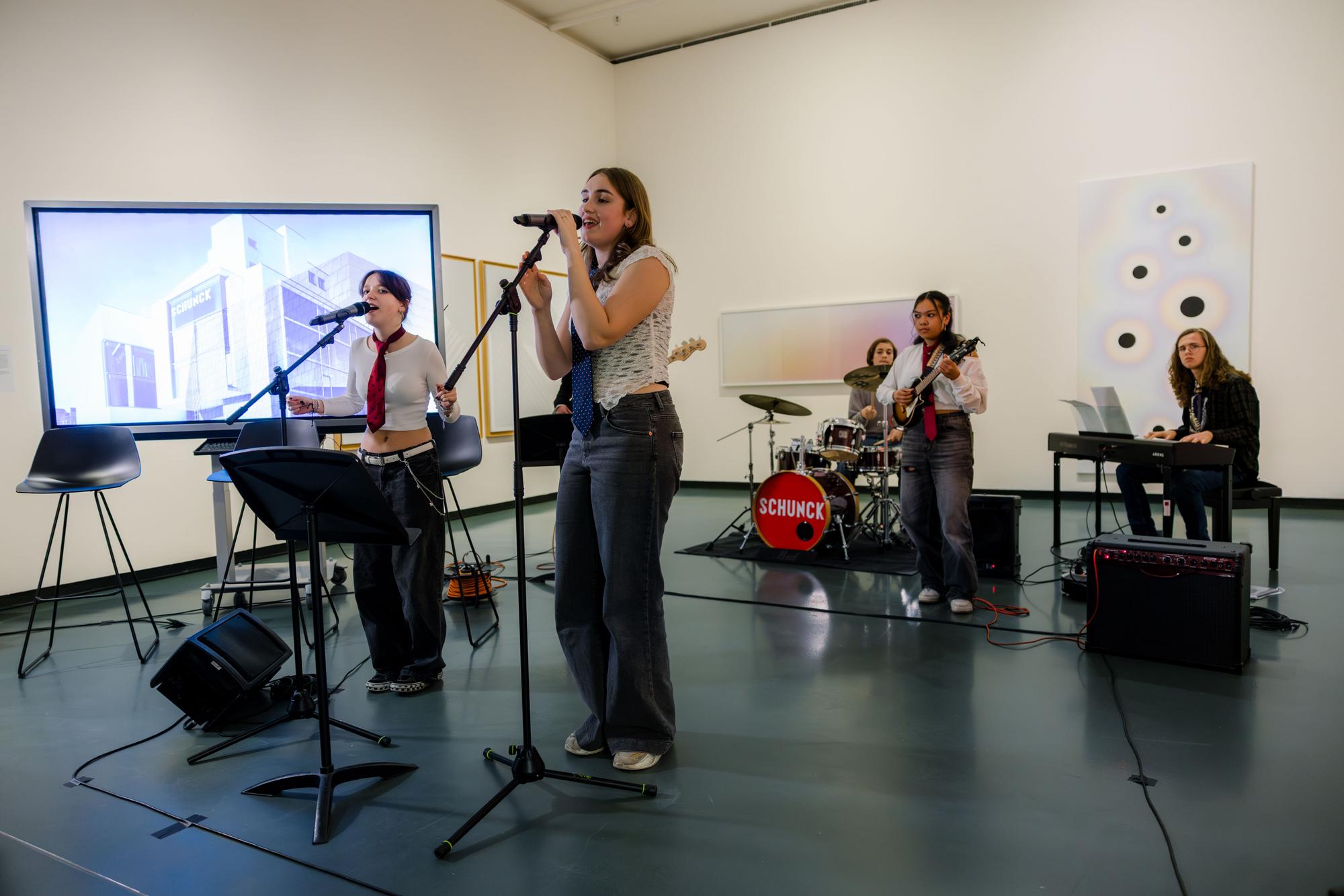 SCHUNCK Academy, jonge band treedt op in een museumruimte met zang, gitaar, drums en keyboard, tussen kunstwerken en een projectiescherm.