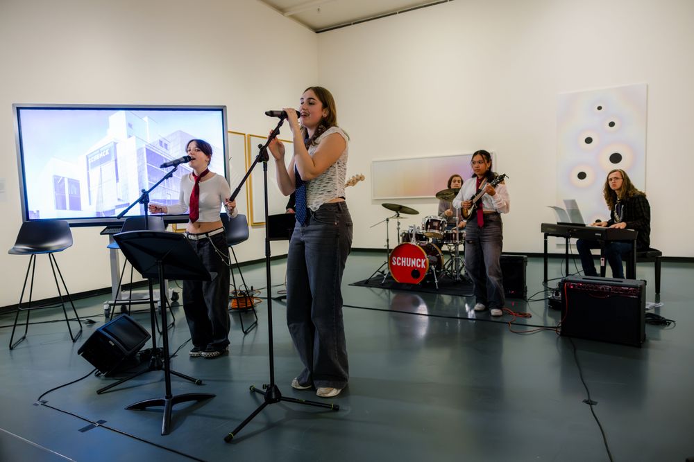 SCHUNCK Academy, jonge band treedt op in een museumruimte met zang, gitaar, drums en keyboard, tussen kunstwerken en een projectiescherm.
