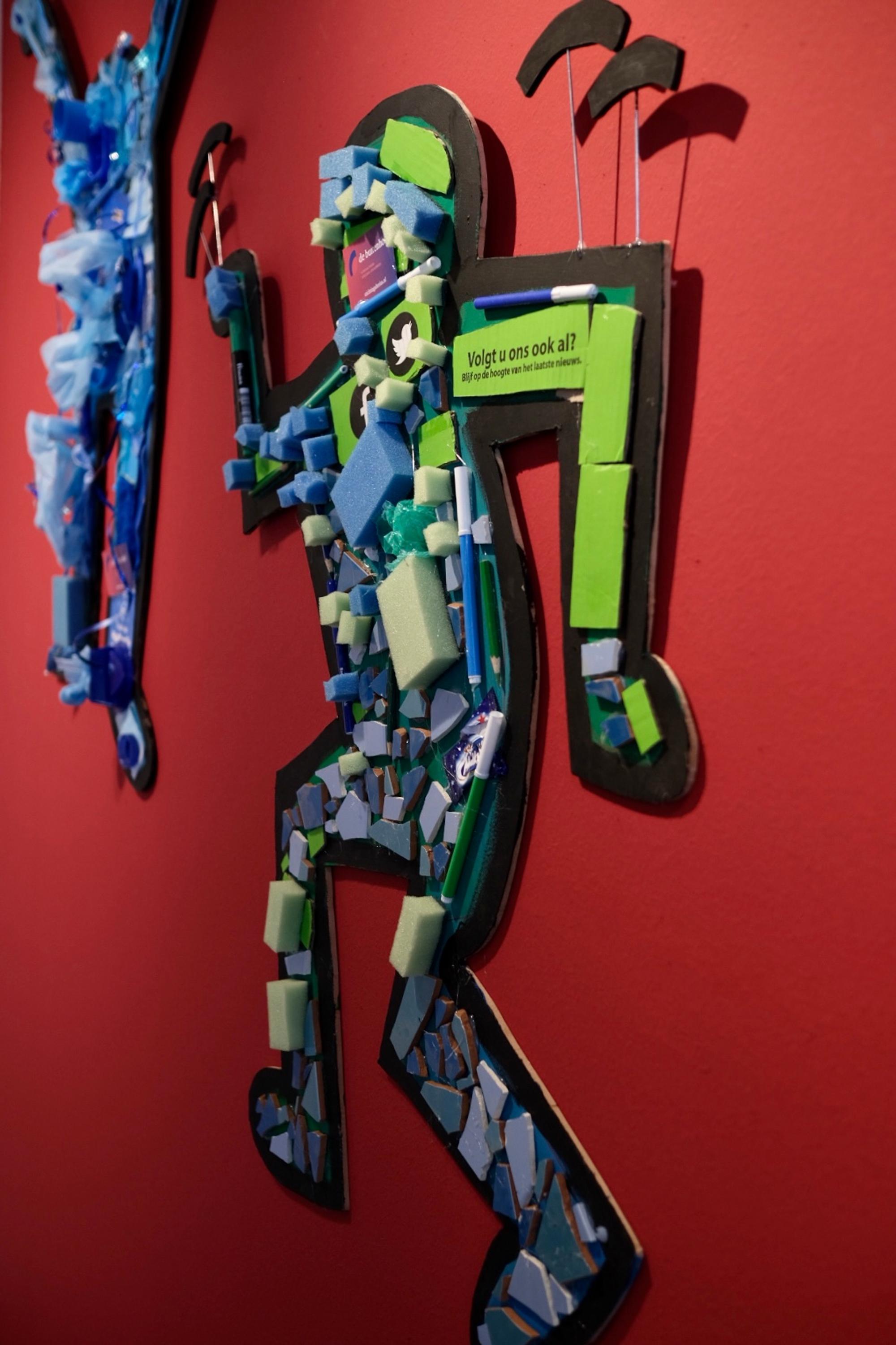 Kunstwerk in de stijl van Keith Haring, gemaakt van schuim, plastic en andere materialen, bevestigd op een rode muur. De figuur is opgebouwd uit gerecyclede onderdelen, waaronder social media-iconen en een tekststicker met de boodschap: “Volgt u ons ook al?”