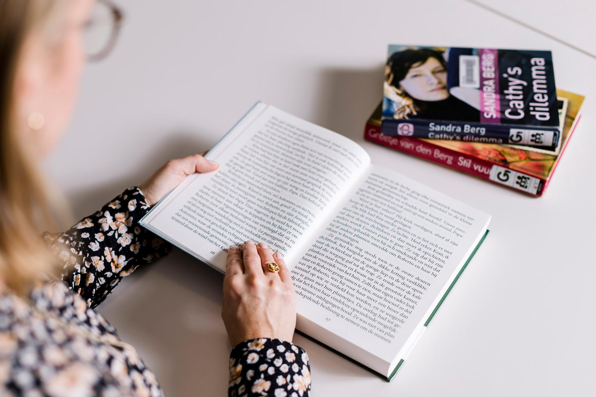 Een vrouw leest een boek. Ze draagt een gouden ring en is gefotografeerd van achteren, over haar schouder, waardoor haar gezicht niet zichtbaar is. In de rechterhoek liggen twee andere boeken.