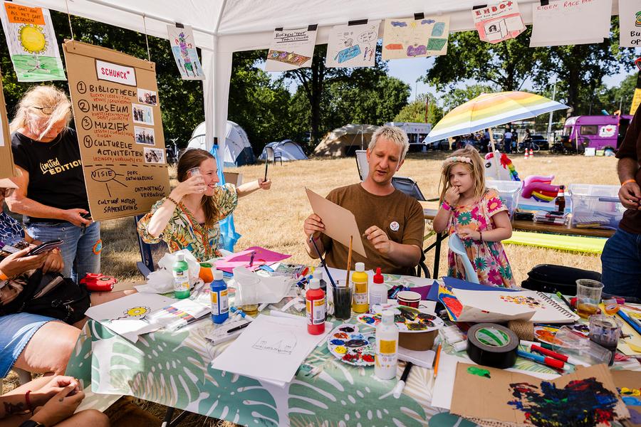 Maarten Bel bekijkt een ontwerp aan een tafel vol verf en tekeningen, omringd door bewoners van de Buurtcamping.
