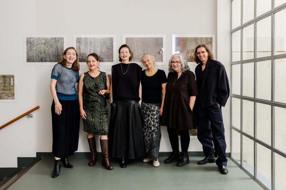 Groepsportret van zes kunstenaars en makers in SCHUNCK, staand voor een wand met fotografische werken tijdens een tentoonstelling.