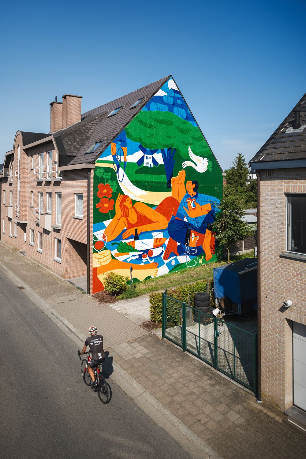 Grote kleurrijke muurschildering van kunstenaar Joren Joshua op de zijgevel van een woongebouw, gezien vanaf de straat met een fietser op de voorgrond.