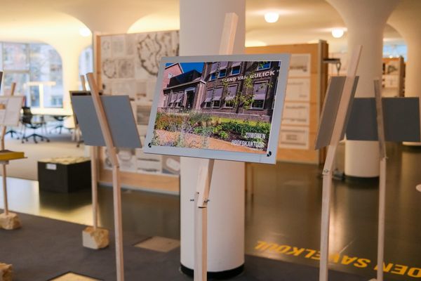 Panelen op houten standaards, waarop foto’s en ontwerpen van gebouwen worden gepresenteerd. Op de voorgrond is een bord te zien met een afbeelding van een oud kantoorgebouw en de tekst “Zó wonen hoofdkantoor.”