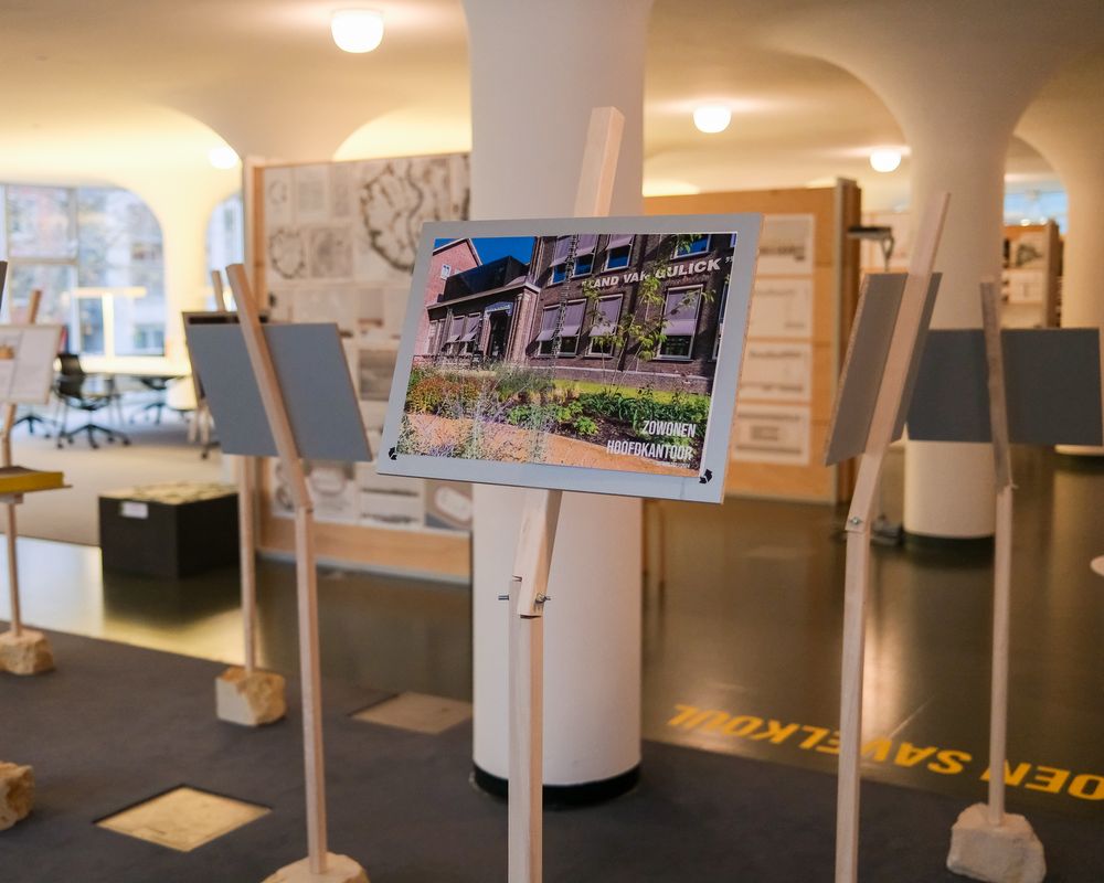 Panelen op houten standaards, waarop foto’s en ontwerpen van gebouwen worden gepresenteerd. Op de voorgrond is een bord te zien met een afbeelding van een oud kantoorgebouw en de tekst “Zó wonen hoofdkantoor.”