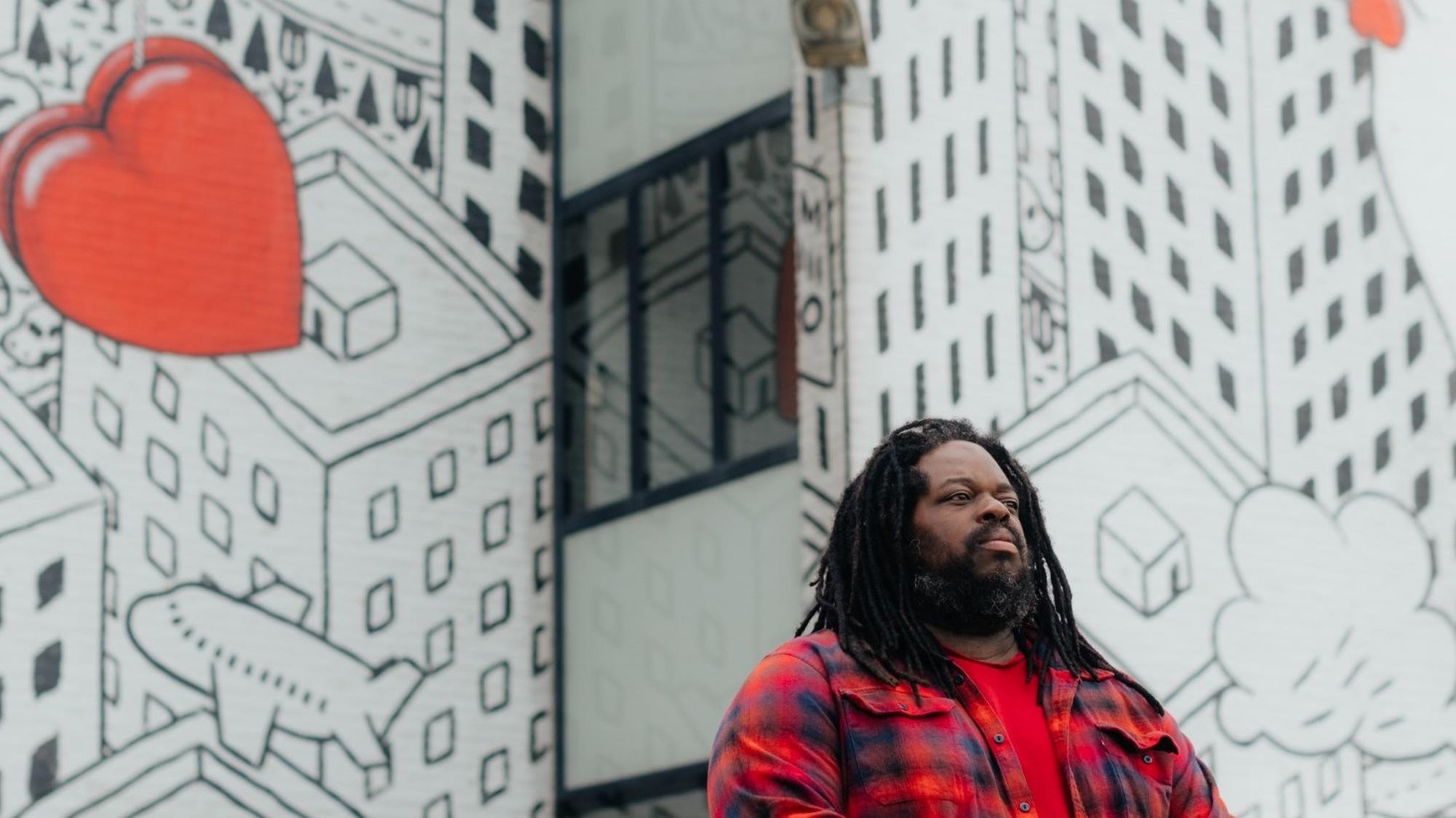 Portret van Raenys Martis met gekruiste armen in een rood geruit overhemd, poserend voor een grote muurschildering met stadsdecor en een rood hart.