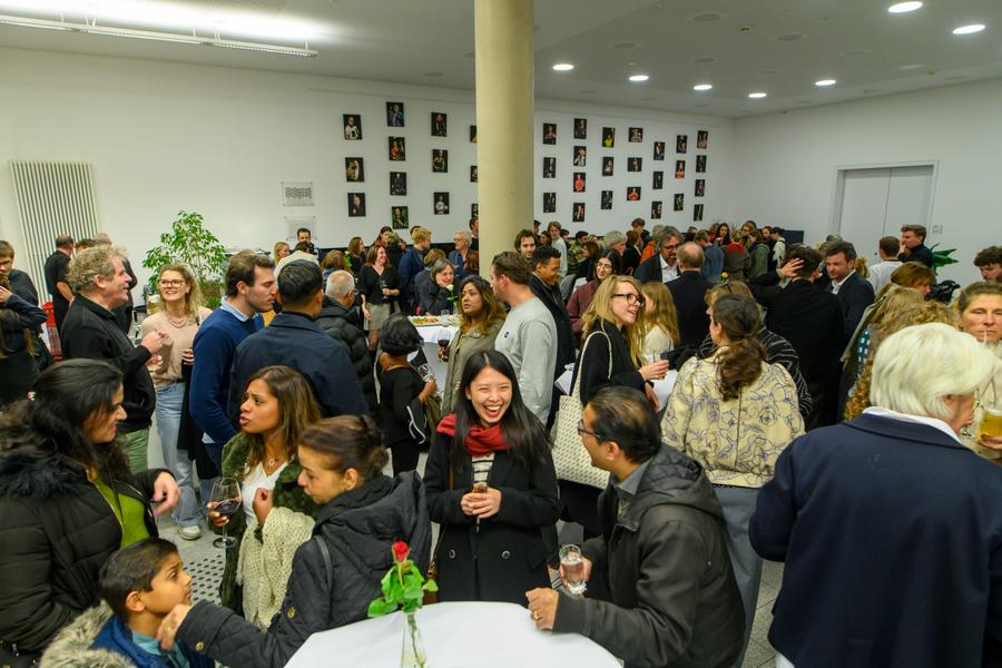Drukke receptie of borrel met mensen die staan en praten in een witte ruimte; op de muur hangen veel kleine portretfoto’s.