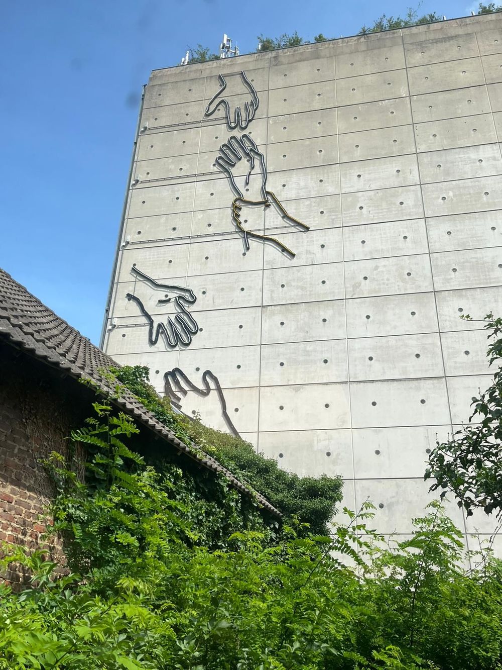 Neonkunstwerk ‘Op handen dragen’ van Chaim van Luit op de betonnen gevel van parkeergarage De Putgraaf in Heerlen, met contouren van grote handen die elkaar vasthouden, gezien vanaf een met groen begroeide binnenplaats.