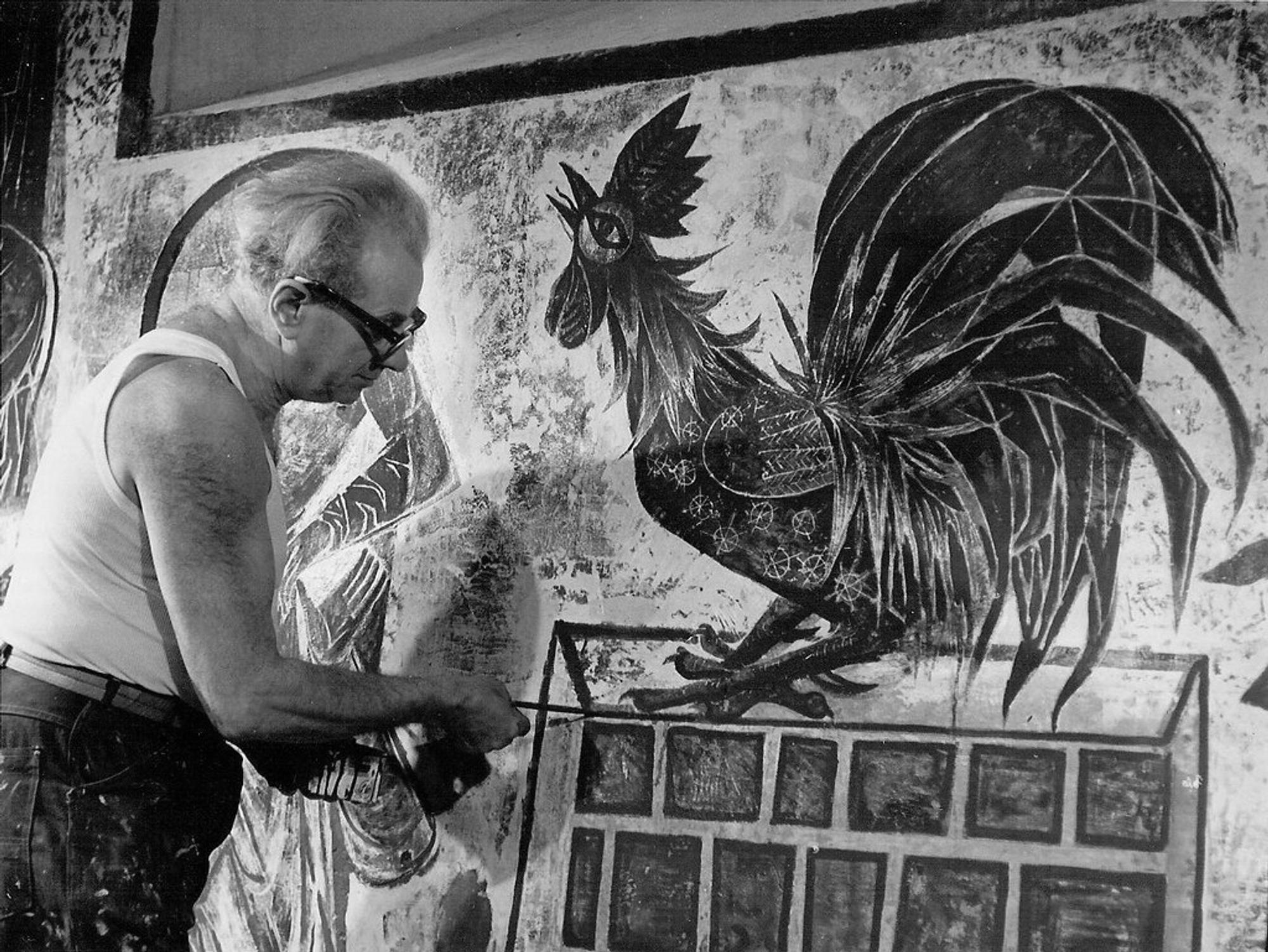 Charles Eyck aan het werk aan muurschildering
Zwart-witfoto van kunstenaar Charles Eyck, met bril en hemd, schilderend aan een muurschildering van een grote haan met sierlijke veren.
