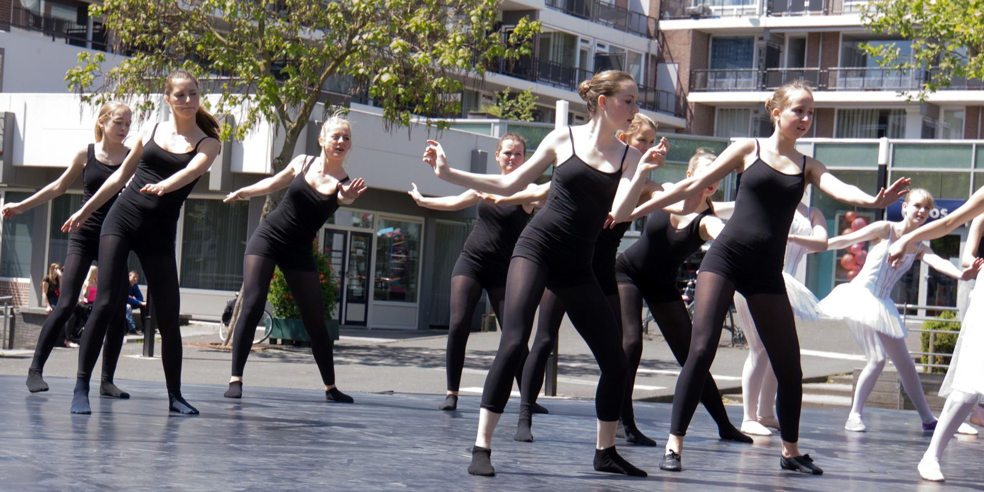 Groep dansers voert een choreografie uit op een buitenpodium, gekleed in zwarte outfits. Op de achtergrond zijn moderne gebouwen en bomen te zien.
