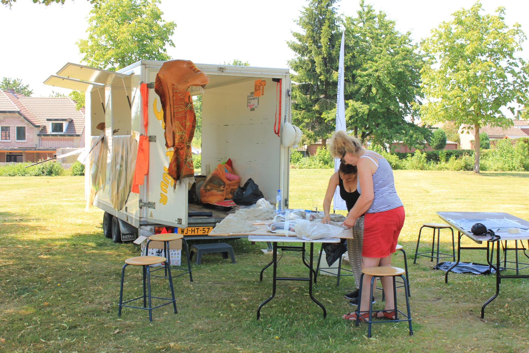 Twee vrouwen werken samen aan een kunstproject aan een tafel in de buitenlucht, naast een open aanhanger met kleurrijke materialen. Op de achtergrond staan bomen en huizen onder een stralend blauwe lucht.