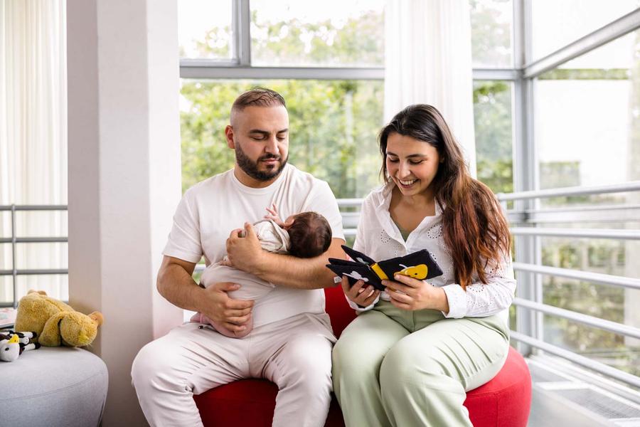 Ouder zit samen met een baby op een rode zitbank terwijl een interactief stoffen boekje wordt voorgelezen. De baby wordt op schoot gehouden en kijkt naar het boekje. 