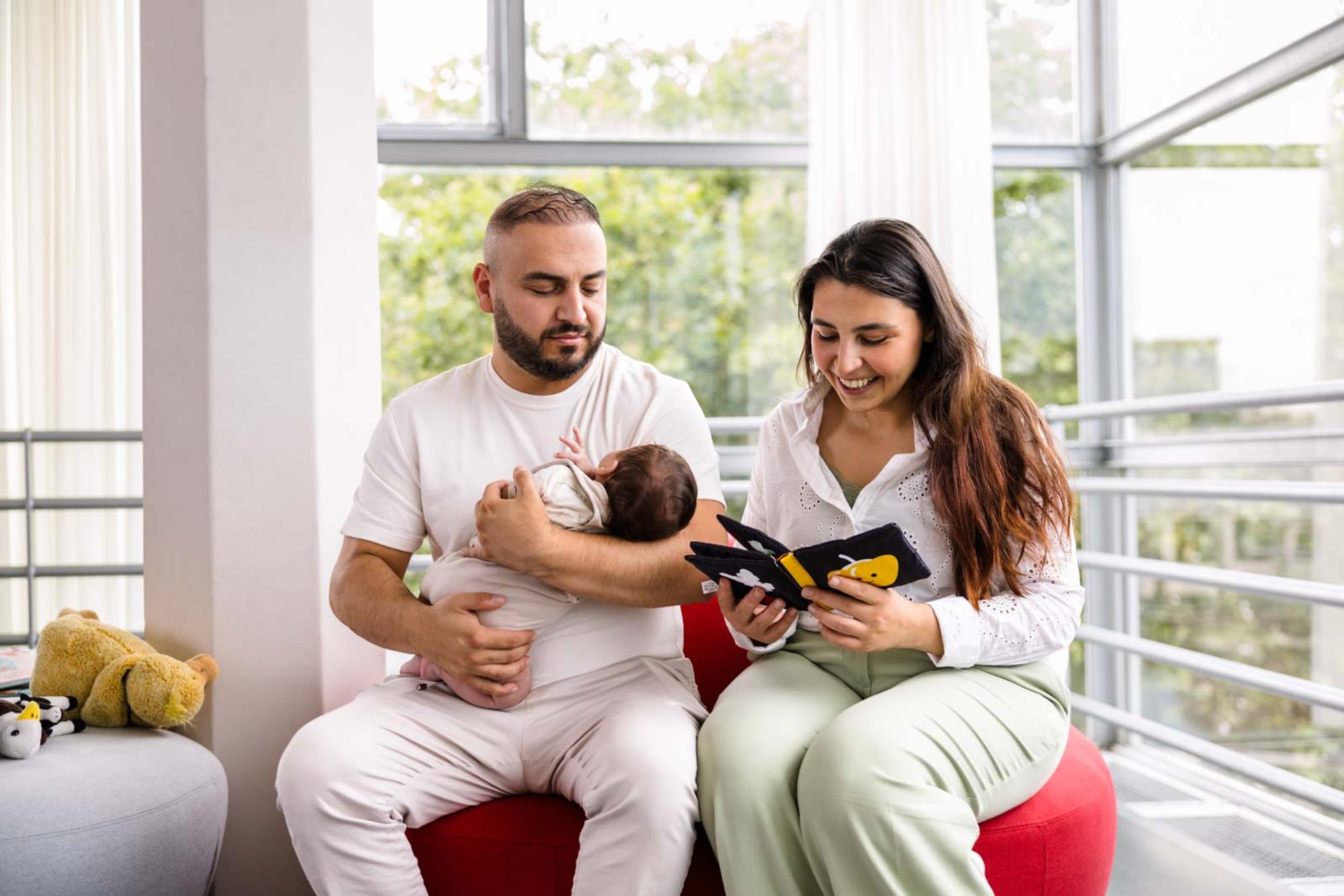 Ouder zit samen met een baby op een rode zitbank terwijl een interactief stoffen boekje wordt voorgelezen. De baby wordt op schoot gehouden en kijkt naar het boekje. 