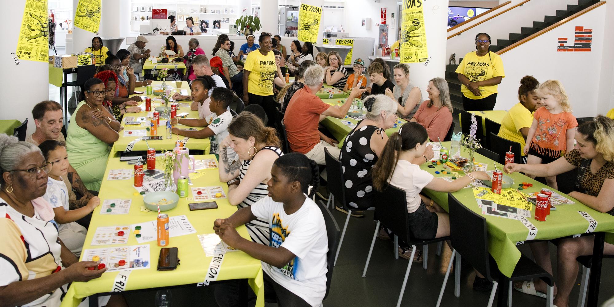 Grote groep mensen zit aan lange tafels in een ruime, lichte zaal waar een spelactiviteit wordt georganiseerd. De tafels zijn bedekt met felgroene tafelkleden en liggen vol met kaartjes, fiches en drankjes. Aan de muren hangen gele posters, en op de achtergrond zijn hoge witte zuilen en een trap zichtbaar.