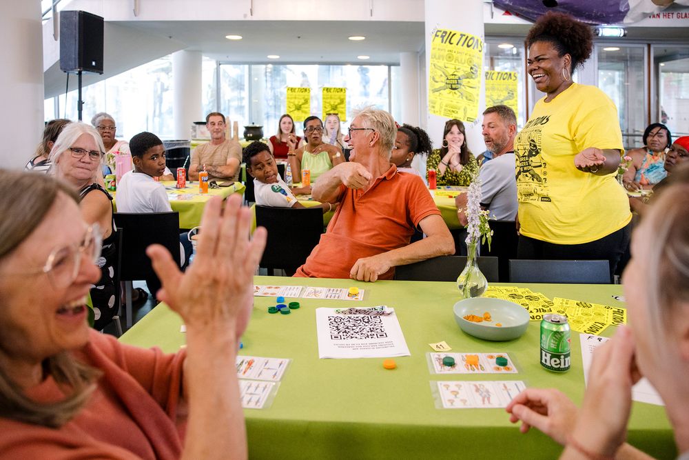 Mensen van verschillende leeftijden zitten aan tafels met groene tafelkleden en spelen enthousiast het spel Bon Ku Ne tijdens Frictions Festival #9; een vrouw in een geel T‑shirt staat lachend tussen de deelnemers, terwijl anderen klappen, lachen en met elkaar praten in een lichte zaal vol gele posters en vrolijke sfeer.