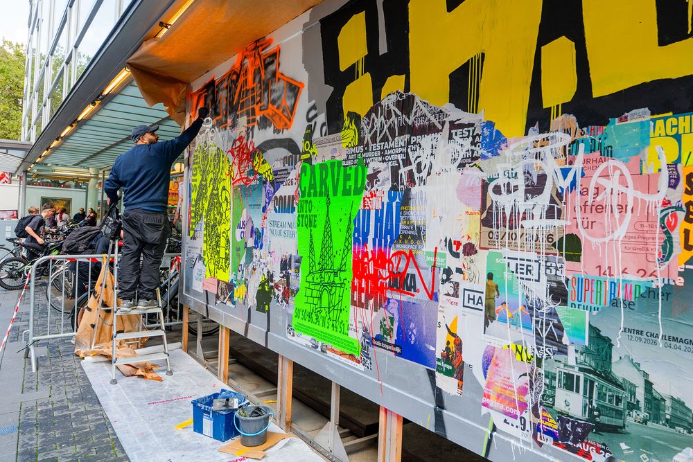 Kunstenaar Massoud Sakhi werkt vanaf een trap aan MODULE #6 bij het Glaspaleis in Heerlen. Het kleurrijke kunstwerk bestaat uit lagen posters, graffiti en grote letters in geel, groen, oranje en zwart.