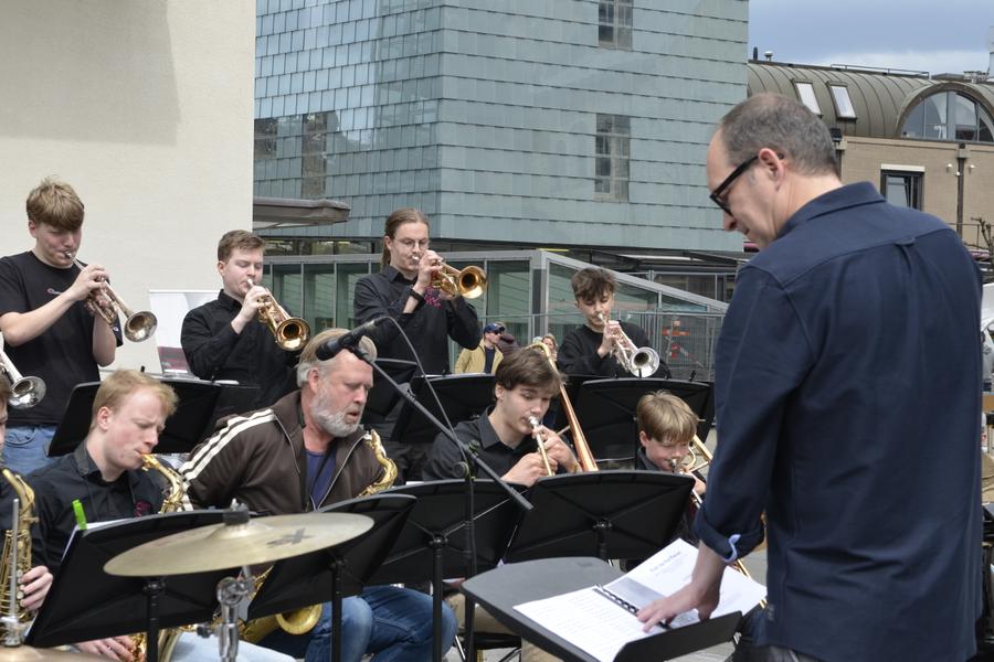 Blaasorkest speelt buiten met muziekstandaards en koperinstrumenten, terwijl een dirigent voor de groep staat. Op de achtergrond zijn stedelijke gebouwen te zien.