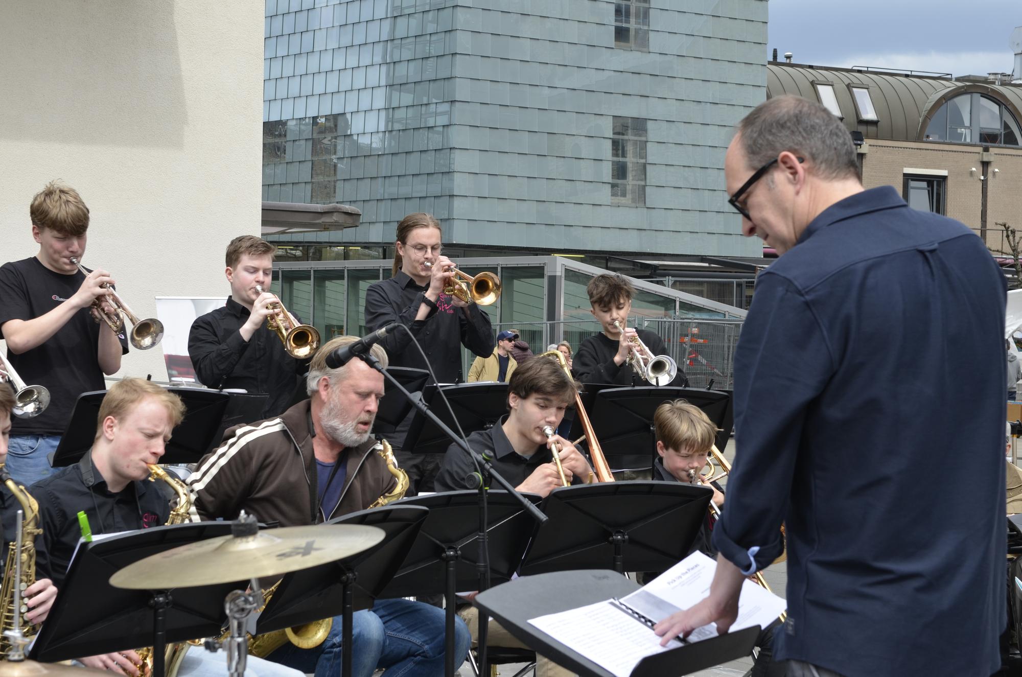 Blaasorkest speelt buiten met muziekstandaards en koperinstrumenten, terwijl een dirigent voor de groep staat. Op de achtergrond zijn stedelijke gebouwen te zien.