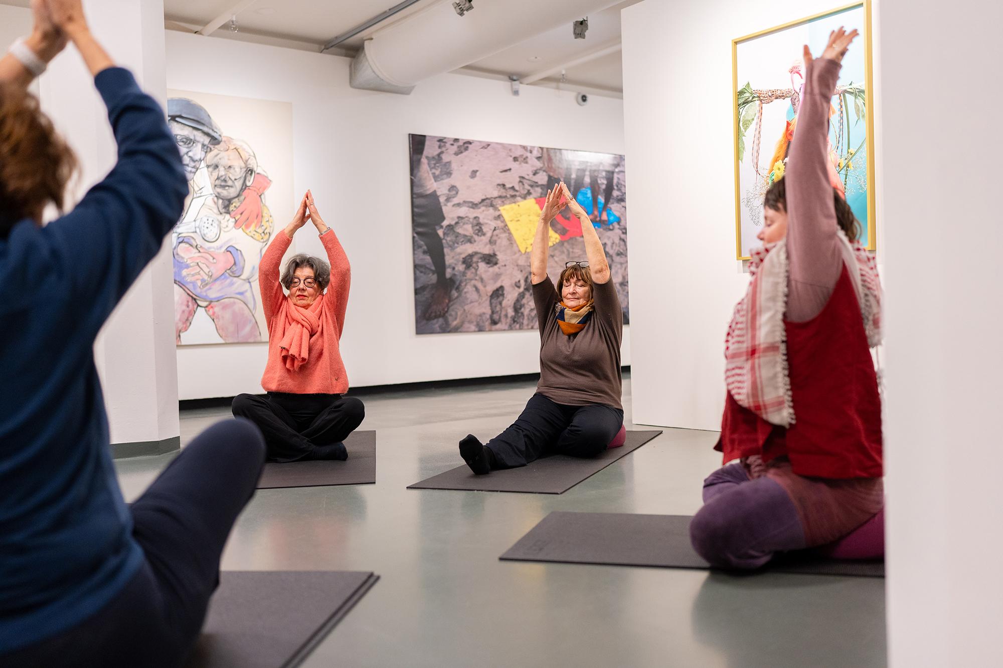 Groep mensen zit op yogamatten in een museumruimte en strekt de armen omhoog tijdens een begeleide oefening, met kunstwerken aan de muren.