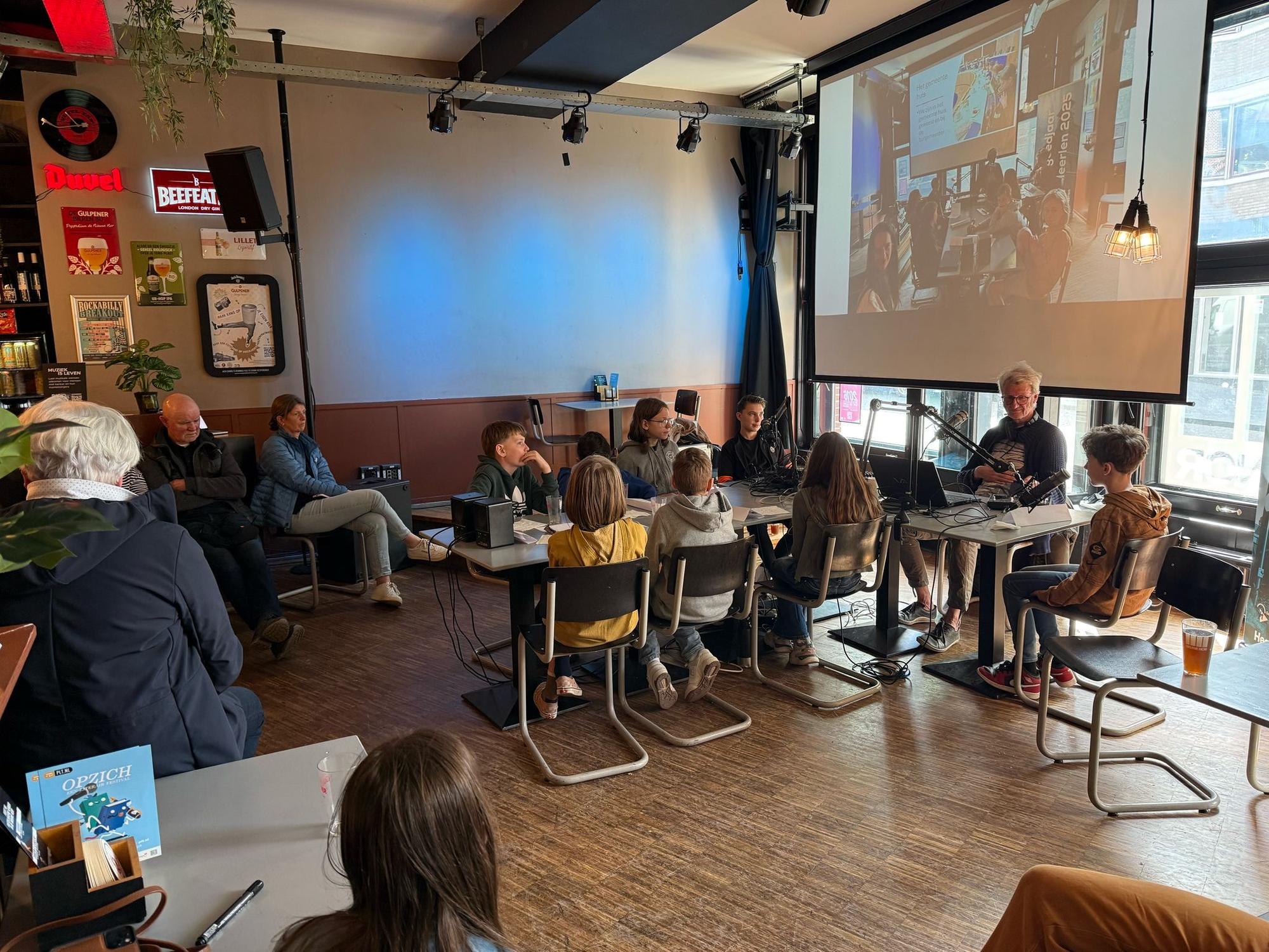 Jongeren zitten aan tafels in een caféachtige ruimte terwijl ze deelnemen aan een opname of podcast, met een begeleider die een microfoon gebruikt en een groot scherm achter hen toont wat er op de opname te zien is. Enkele volwassenen kijken vanaf de zijkant toe.