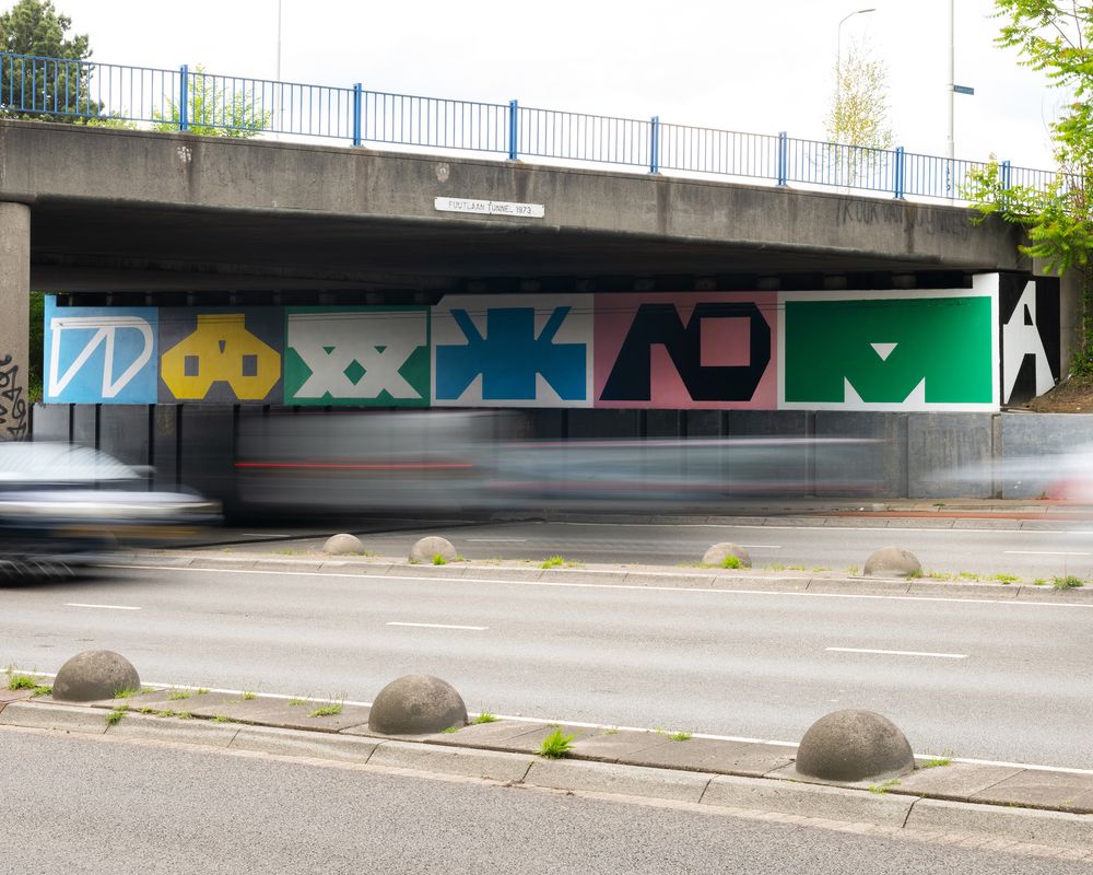 Grote, kleurrijke muurschildering met abstracte letters onder een viaduct; op de voorgrond rijdt verkeer met bewegingsonscherpte over de weg.