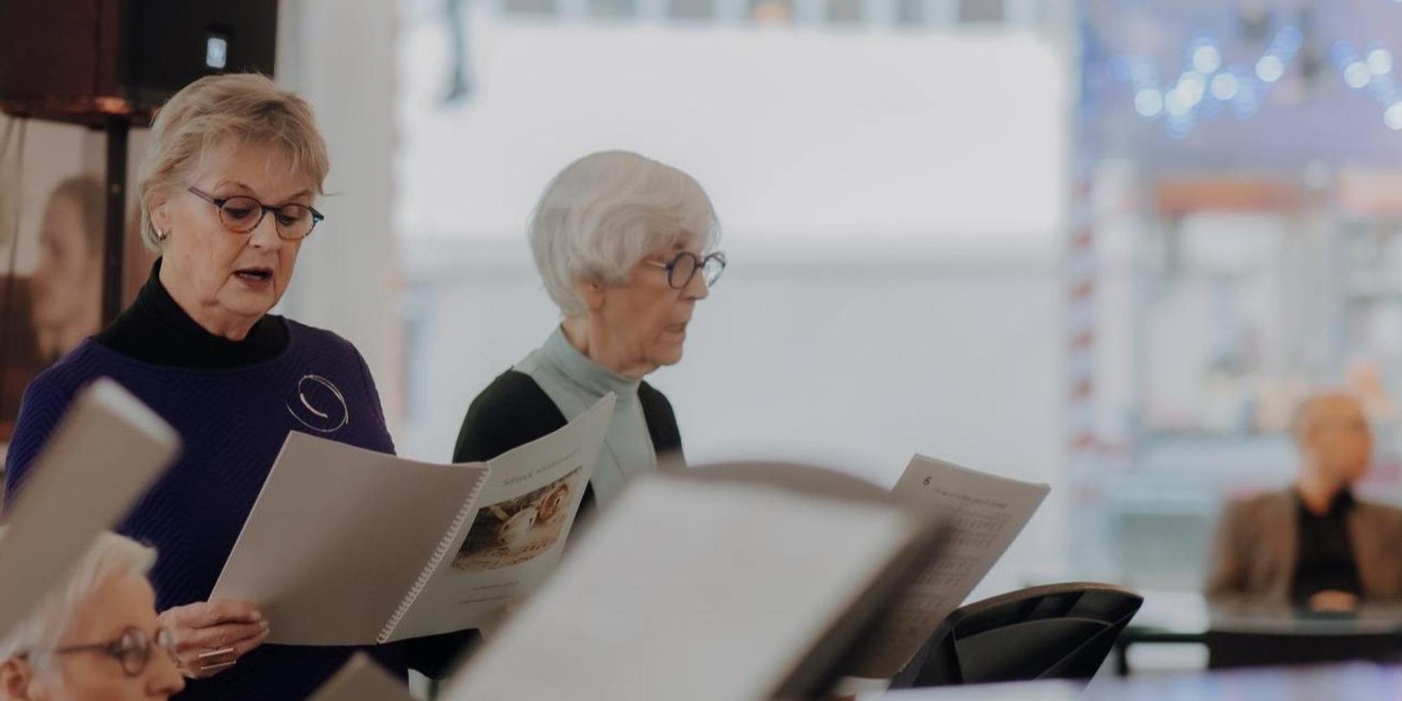Een groep oudere zangers staat samen en zingt van bladmuziek tijdens een repetitie of optreden. De focus ligt op concentratie en samenzang in een lichte, openbare ruimte.