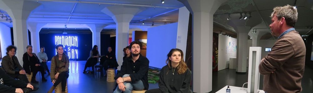 Leden van de Badeklasse zitten op krukken in de museumzaal, voor hen staat kunstenaar David Bade. Op de achtergrond is het kunstwerk 'Patriarchy is History' te zien, gemaakt van blauwe neonbuizen.