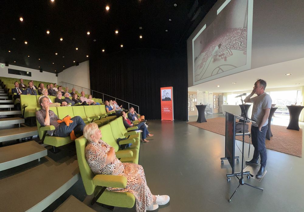 Een spreker staat bij een katheder voor een groot projectiescherm in een moderne zaal met groene stoelen, waarin een aandachtig publiek zit te luisteren naar een presentatie over Keith Haring.