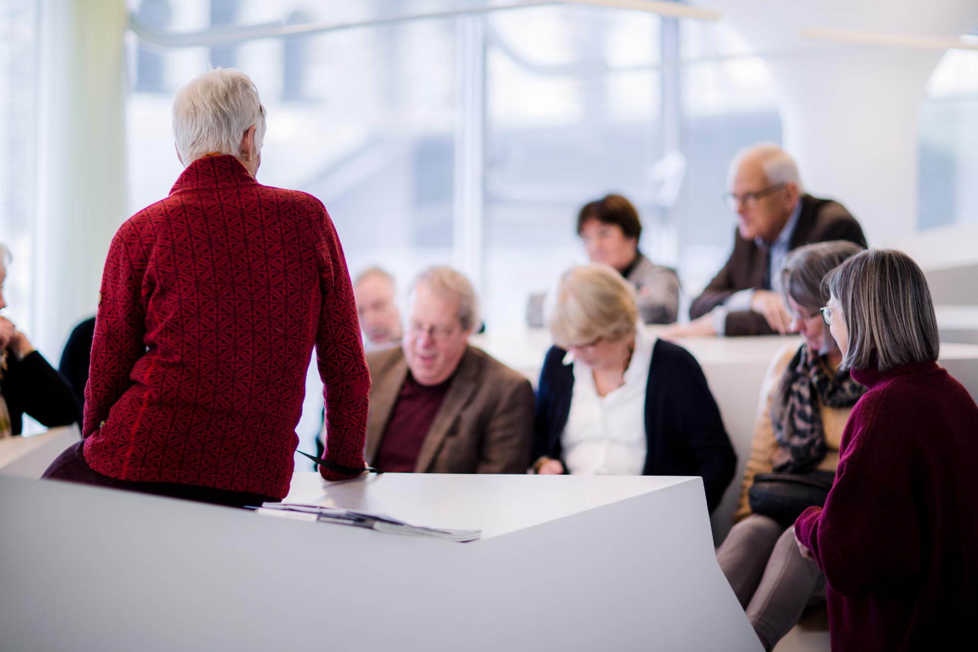 Een groep senioren luistert aandachtig naar een spreker in een moderne, lichte ruimte, vermoedelijk tijdens een lezing of bijeenkomst.